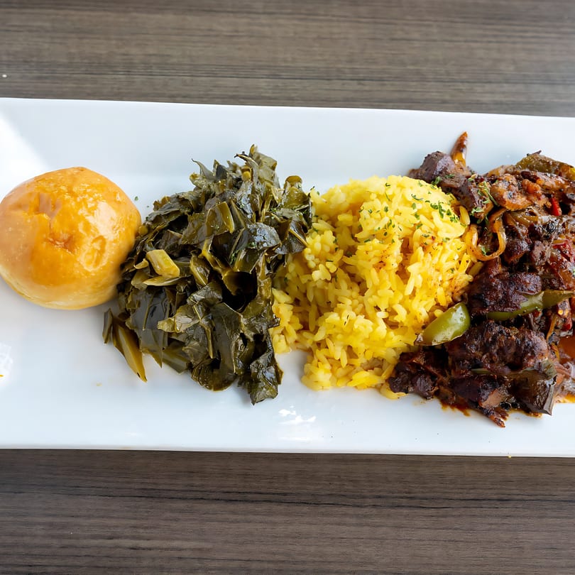A soul food plate with a golden roll, collard greens, yellow rice, and stewed meat.