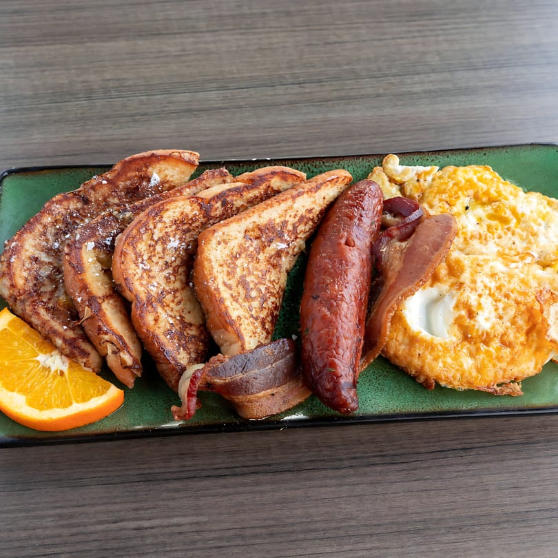 A soul food breakfast plate with French toast, sausage, bacon, scrambled eggs, and an orange slice.