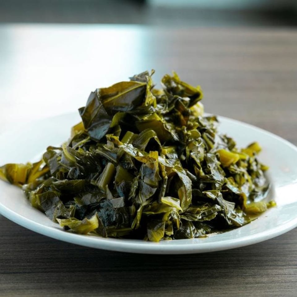A white plate filled with a generous serving of dark green, cooked collard greens.