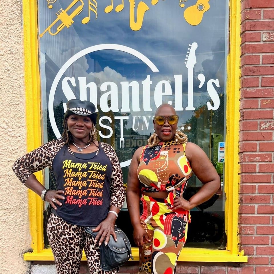 Two women in vibrant outfits pose in front of Shantell's Just Until Restaurant & Lounge.