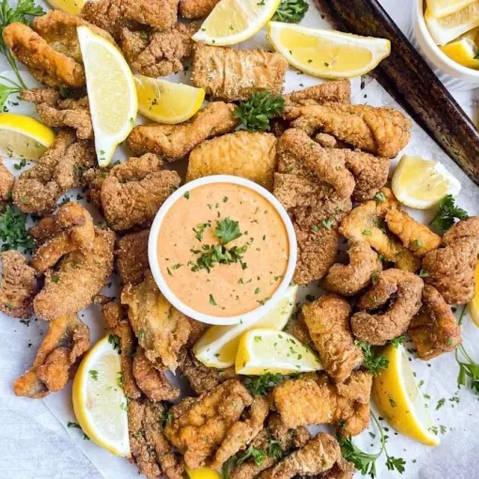 Crispy fried catfish pieces with lemon wedges and a side of creamy dipping sauce, garnished with parsley.