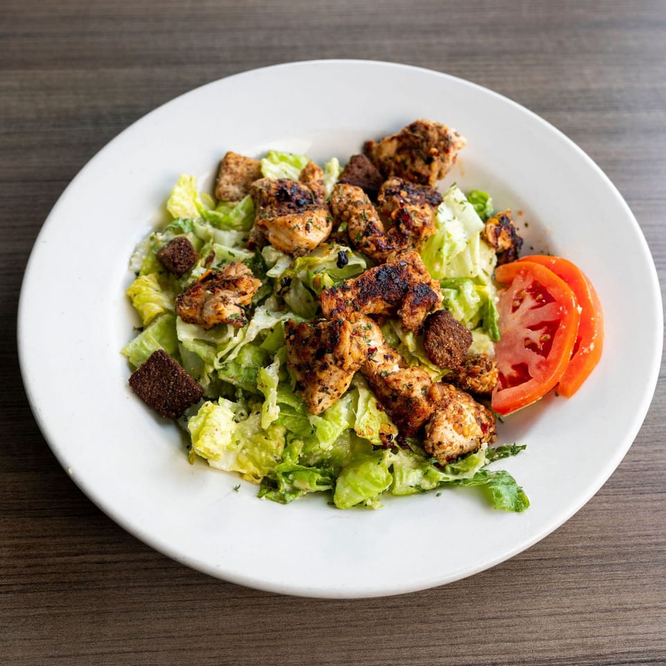 A Caesar salad with grilled chicken, croutons, and tomato slices on a white plate.