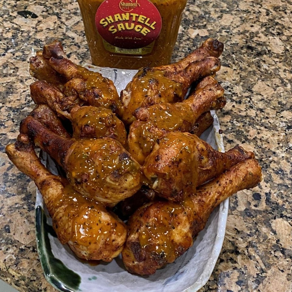 A platter of golden-brown chicken drumsticks, generously coated in Shantell's signature sauce, on a granite countertop.